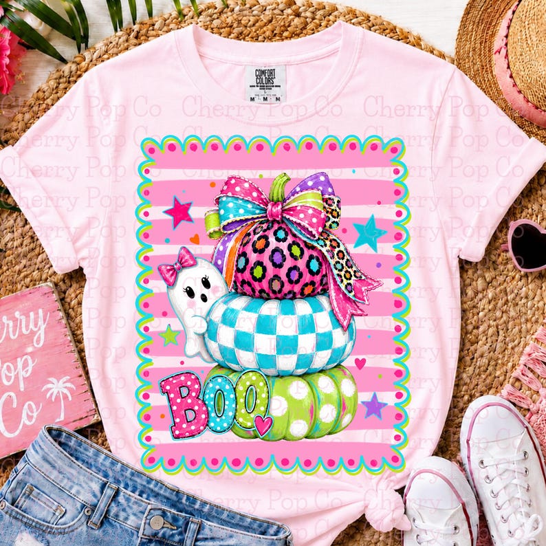 May include: A light pink short-sleeve t-shirt featuring a graphic of three stacked pumpkins with a large colorful bow on top. The pumpkins have leopard print, teal checkered, and green polka dot patterns. A small white ghost and the text BOO are included.