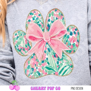 Puede incluir: Sudadera gris claro con un dise&ntilde;o de gran huella de pata. La huella est&aacute; llena de un estampado floral en rosa, verde y azul, delineado con purpurina dorada. Un gran lazo rosa est&aacute; centrado en la huella. El texto "CHERRY POP CO" y "PNG DESIGN" est&aacute;n en la parte inferior.