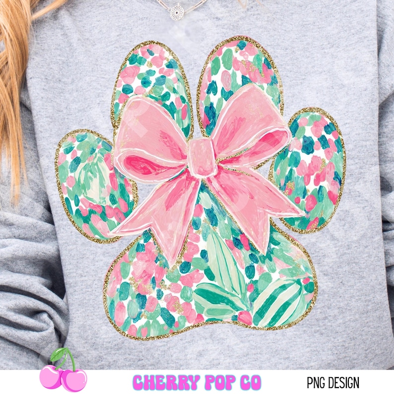Puede incluir: Sudadera gris claro con un dise&ntilde;o de gran huella de pata. La huella est&aacute; llena de un estampado floral en rosa, verde y azul, delineado con purpurina dorada. Un gran lazo rosa est&aacute; centrado en la huella. El texto "CHERRY POP CO" y "PNG DESIGN" est&aacute;n en la parte inferior.