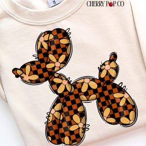 以下が含まれることがあります： クリーム色のスウェットシャツ。風変わりなバルーン犬のデザインが特徴です。犬は黒とオレンジの市松模様で装飾され、黄色のデイジーの花とピンクの中心がアクセントになっています。「CHERRY POP CO」の文字が上部に印刷されています。