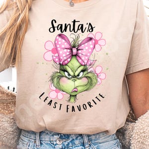 Puede incluir: Camiseta beige con un dise&ntilde;o del Grinch, un lazo rosa con lunares y flores rosas. El texto "Santa's Least Favorite" est&aacute; impreso encima y debajo de la cara del Grinch en escritura negra.