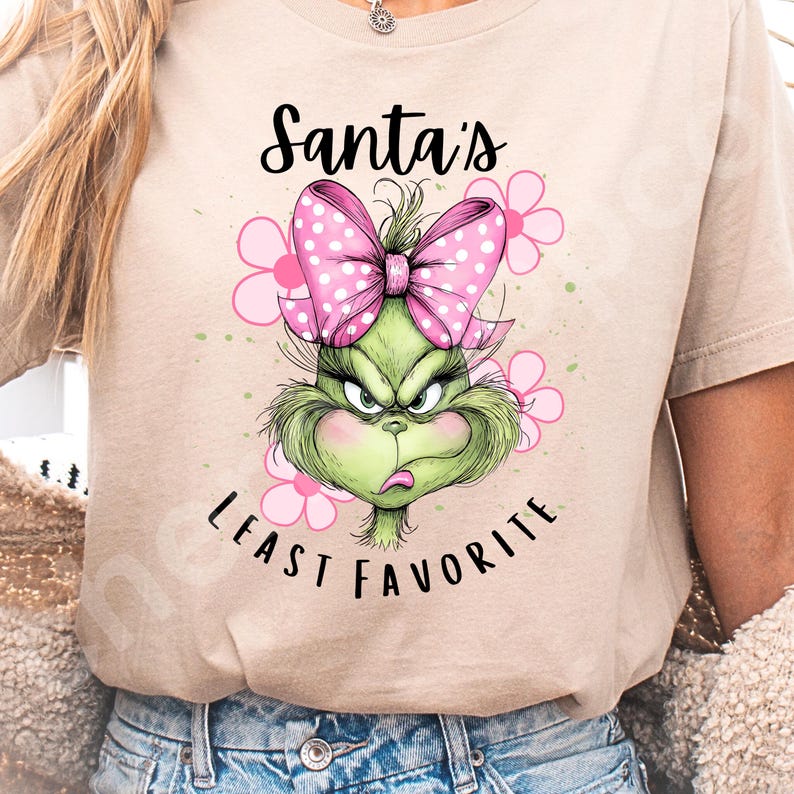 Puede incluir: Camiseta beige con un dise&ntilde;o del Grinch, un lazo rosa con lunares y flores rosas. El texto "Santa's Least Favorite" est&aacute; impreso encima y debajo de la cara del Grinch en escritura negra.