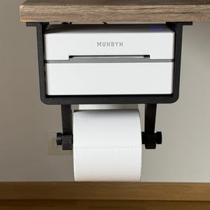 May include: A white label printer with the text "MUNBYN" on the front, mounted under a wooden desk with a black metal bracket. A roll of white paper is attached to the bracket.