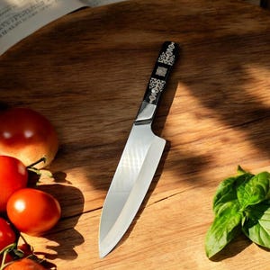 Peut inclure: Un couteau de cuisine argenté avec un manche noir orné de motifs argentés complexes, posé sur une planche à découper en bois avec des tomates et du basilic.