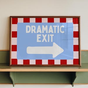 May include: A framed sign with the words "DRAMATIC EXIT" and a white arrow on a light blue background. The frame has a red and white checkered border. The sign rests on a green shelf.