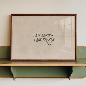 May include: A framed print with the text "I SEE LONDON I SEE FRANCE" rests on a green and wood-toned shelf. The print has a brown frame and a light-coloured background. The shelf is mounted on a white wall.
