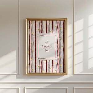 Op de afbeelding: Een ingelijste kunstdruk met een gouden lijst. Het kunstwerk heeft een witte achtergrond met een rood-wit gestreept patroon en kleine gele hartjes. Het midden heeft een witte rechthoek met de woorden "ok love you, bye".