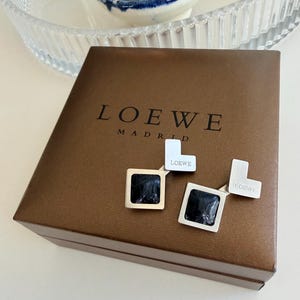 Può includere: Un paio di orecchini quadrati con centri neri e montature argentate e dorate, appoggiati su una scatola marrone. La scatola è impressa con la parola "LOEWE" sopra "MADRID". Gli orecchini hanno un design geometrico.