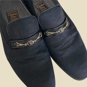 Könnte beinhalten: Ein Paar schwarze Leder-Slipper mit goldenen Schnallen und dem Schriftzug "Christian Dior" auf der Oberseite jedes Schuhs.