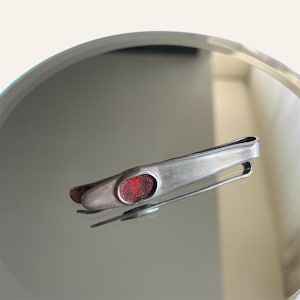 Pode incluir: Um prendedor de gravata prateado com uma inserção vermelha retangular. O prendedor está sobre uma superfície reflexiva, mostrando seu design elegante e o contraste com o detalhe vermelho.