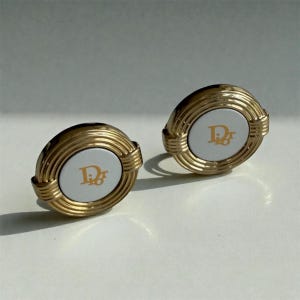 May include: A pair of gold-toned oval earrings. Each earring features a white center with the letters "Dior" in gold. The earrings have a ridged gold border and a post backing. The earrings are displayed on a white surface.