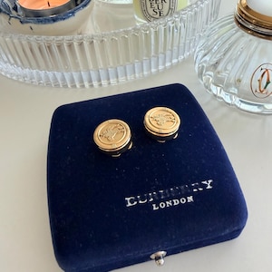 Può includere: Un paio di gemelli dorati con un design circolare, appoggiati su un cofanetto di velluto blu scuro. Il cofanetto reca la scritta "BURBERRY LONDON" in lettere bianche. I gemelli presentano un motivo inciso dettagliato.