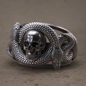 May include: A silver ring featuring a skull encircled by two snakes. The ring has a detailed, textured design, with the snakes' scales and the skull's features clearly visible. The ring is a gothic or biker-style accessory.