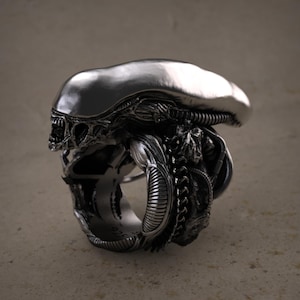 Puede incluir: Un anillo plateado detallado que representa la cabeza de una criatura Alien. El anillo muestra la distintiva cabeza alargada de la criatura, su exoesqueleto texturizado y sus amenazantes rasgos faciales. El anillo es una joya única.