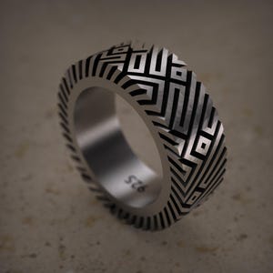 May include: A silver ring with a geometric pattern. The ring has a repeating design of black and silver lines, creating a maze-like effect. The band is thick and has a textured edge. The ring is marked with "925".