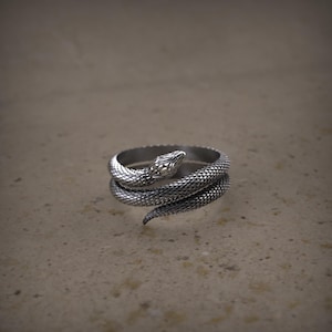 K&ouml;nnte beinhalten: Ein silberner Schlangenring mit einem detaillierten Kopf und Schuppen. Die Schlange ist um sich selbst gewickelt und bildet einen Kreis.