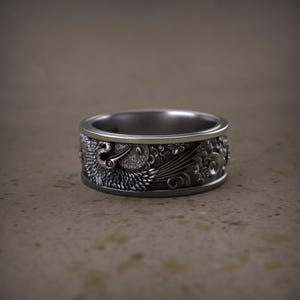 May include: A silver ring with detailed relief carvings. The ring features a bird design with floral and wave patterns. The ring is a wide band style, with a polished interior and a matte finish on the exterior.
