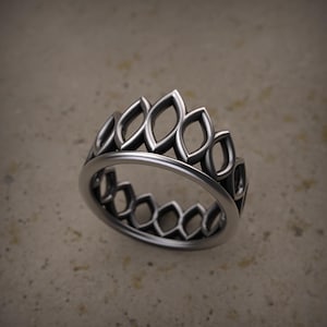 Puede incluir: Un anillo plateado con un diseño repetitivo en forma de hoja. El anillo presenta detalles calados a lo largo de la banda y una serie de formas alargadas y puntiagudas alrededor de la parte superior, creando una apariencia de corona. El anillo está sobre un fondo neutro.