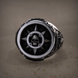 May include: A silver ring with a detailed design. The ring features a central black circular emblem with a skull, a starburst pattern, and a clear gem. The ring's band has intricate detailing.