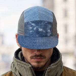 May include: A five-panel cap featuring a patchwork design of blue denim and grey fabric, paired with a jacket that has a brown suede upper section, olive green nylon hood and collar, and a front zip closure with two dark drawstrings.