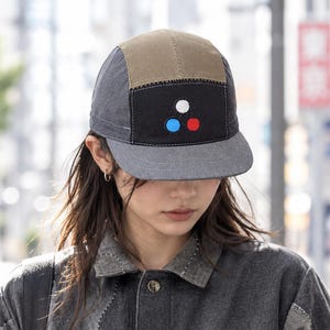 Puede incluir: Una gorra de cinco paneles con visera y laterales gris carbón, panel superior beige y panel frontal negro con tres círculos bordados en blanco, azul y rojo. La gorra se combina con una chaqueta abotonada hecha de parches de mezclilla gris.