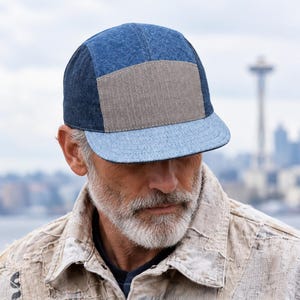 Könnte beinhalten: Eine Fünf-Panel-Kappe mit Color-Block-Design, bestehend aus dunkelblauen Denim-Seitenteilen, einem mittigen hellbraunen rechteckigen Panel und einem flachen Schirm aus hellblauem Denim, die durch sichtbare Nähte miteinander verbunden sind.