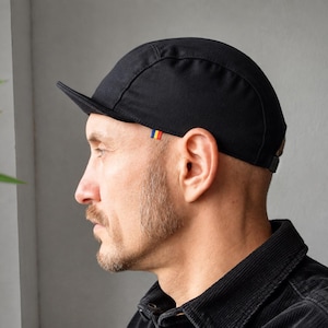 May include: A solid black cycling cap with a short, stiffened brim and a small, rectangular woven tag featuring blue, yellow, and red vertical stripes positioned on the side near the temple area, providing a clean and functional headwear design.