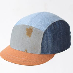Peut inclure: Casquette à cinq panneaux avec un design patchwork. Le chapeau présente un dessus en jean bleu clair, un panneau gris avec une tache brune et un panneau latéral en jean bleu foncé. La visière est orange.