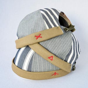 Può includere: Un berretto senza visiera con un design patchwork in tessuto grigio gessato e pannelli a righe blu navy e bianche. Il berretto presenta un bordo arrotolato in tela color cuoio decorato con piccoli simboli ricamati in rosso, tra cui una X, un triangolo e una Y.