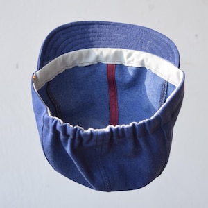 May include: A blue cap with a curved brim and a white interior band. The cap has a red stripe down the center and a gathered elastic band. The cap is made of a soft, durable material.
