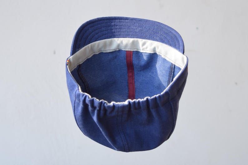 May include: A blue cap with a curved brim and a white interior band. The cap has a red stripe down the center and a gathered elastic band. The cap is made of a soft, durable material.