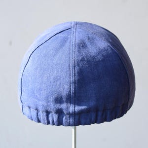 May include: A blue cap with a rounded crown and a stitched seam running down the center. The cap has a ribbed band at the base and is made of a textured fabric. The cap is displayed against a plain background.