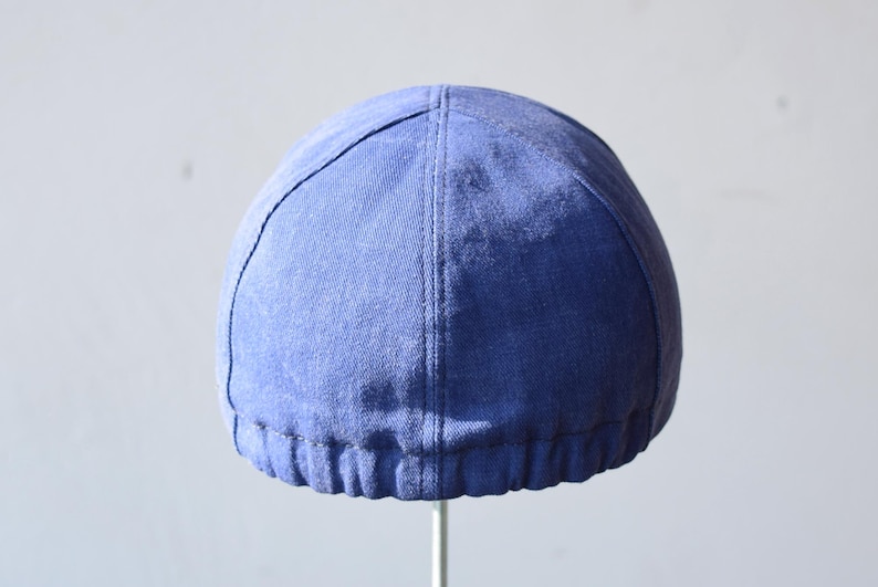 May include: A blue cap with a rounded crown and a stitched seam running down the center. The cap has a ribbed band at the base and is made of a textured fabric. The cap is displayed against a plain background.