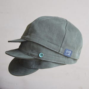 Op de afbeelding: Drie saliegroene petten op elkaar gestapeld. Elke pet heeft een kleine klep en een knoop aan de zijkant. Een kleine blauwe vierkante patch met een wit ontwerp is zichtbaar aan de zijkant van de bovenste pet.