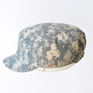 May include: A digital camouflage patrol cap in shades of gray, tan, and white. The cap features a short brim and a small, decorative emblem on the side. The hat is a military-style accessory.