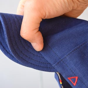 May include: Close-up of a navy blue hat with a curved brim. The hat features a red triangle emblem and detailed stitching. The fabric appears to be a durable cotton or similar material, suitable for outdoor wear.