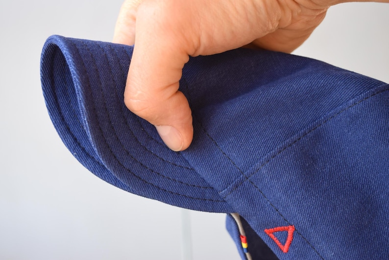 May include: Close-up of a navy blue hat with a curved brim. The hat features a red triangle emblem and detailed stitching. The fabric appears to be a durable cotton or similar material, suitable for outdoor wear.