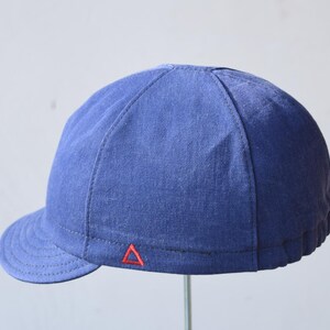May include: A blue cap with a short brim. The cap is made of a textured fabric and features a small red triangle emblem near the brim. The cap is a solid blue color and has a rounded crown.