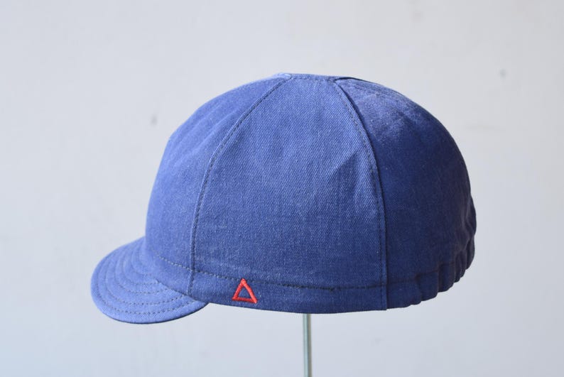 May include: A blue cap with a short brim. The cap is made of a textured fabric and features a small red triangle emblem near the brim. The cap is a solid blue color and has a rounded crown.
