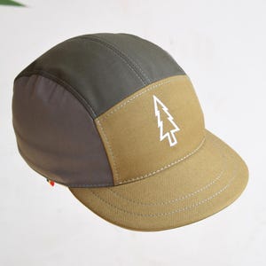 May include: A five-panel cap in grey and tan, with a white tree graphic on the front. The cap has a curved peak and a structured design, suitable for outdoor activities.