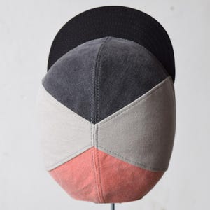 Puede incluir: Una gorra de béisbol de cinco paneles con un diseño de bloques de color, que presenta una visera y una parte superior negras. El cuerpo principal consta de paneles triangulares en gris carbón, gris claro y rosa coral, unidos por costuras visibles.