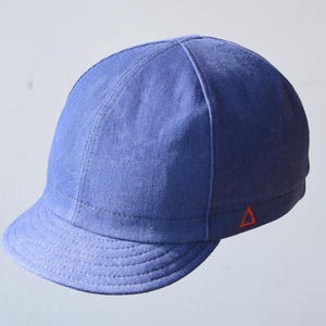 May include: A blue cap with a short brim and a red triangle logo on the side. The cap is made of a textured fabric and has a stitched design. This hat is a stylish accessory.