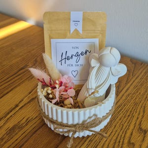Könnte beinhalten: Eine weiße, gerippte Keramikschale mit einer Kordel, gefüllt mit einem braunen Papiertütchen mit der Aufschrift "VON HERZEN FÜR DICH" und einem Herzsymbol, einer weißen Engelsfigur sowie getrockneten rosa und cremefarbenen Blumen und Gräsern.