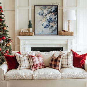 May include: A cozy living room scene featuring a white sofa adorned with red and plaid pillows. A framed artwork of blue and white Christmas ornaments hangs above a fireplace. A decorated Christmas tree is visible on the left.