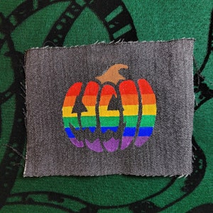 May include: A gray fabric patch with a rainbow colored pumpkin design. The pumpkin has a traditional jack-o-lantern face.