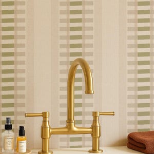 May include: A brushed gold bridge faucet with two lever handles sits above a beige sink. To the left, two small clear bottles with labels rest on a circular tray. To the right, a folded brown towel lies on the counter against a striped wall.