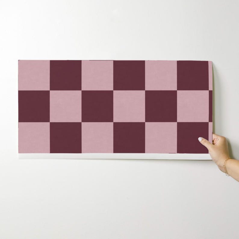 Puede incluir: Un rollo de papel pintado con un dise&ntilde;o de tablero de ajedrez. El dise&ntilde;o alterna cuadrados de color rosa empolvado y borgo&ntilde;a oscuro. El papel pintado est&aacute; sujeto por una mano sobre un fondo blanco.