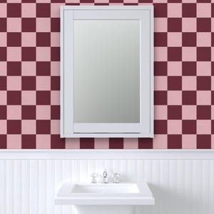 Puede incluir: Un espejo con marco blanco cuelga de una pared con un patr&oacute;n de cuadros en tonos rosa y burdeos. Debajo del espejo hay un lavabo blanco con un grifo cromado, sobre una pared con paneles blancos.