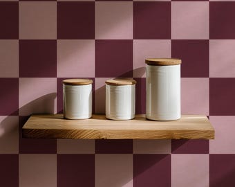 May include: Three white ceramic canisters with wooden lids of varying sizes sit on a wooden shelf. The canisters are cylindrical and have a smooth, glossy finish. The background is a checkered pattern of dark and light pink squares.