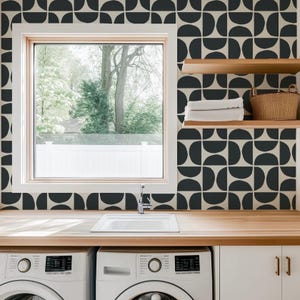 May include: Two white washing machines with a black and white geometric pattern wallpaper behind them. The wallpaper has a repeating pattern of black semi-circles. There is a white countertop with a white sink and a faucet. There are two wooden shelves above the countertop with white towels and a wicker basket on them.